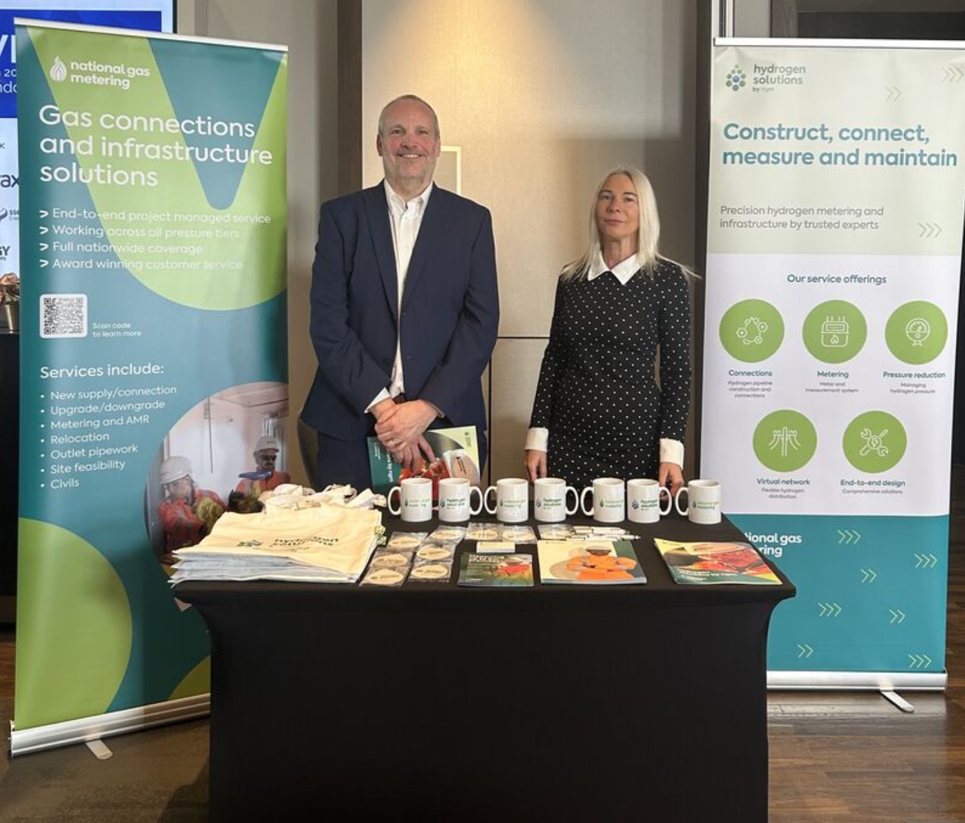 Two people standing at a National Gas Metering and Hydrogen Solutions promotional stand with banners, mugs, and brochures.